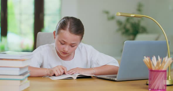 Focused Teen Girl at the Desk Reads a Book Tracking Lines with Finger Laptop Aside Books Notebooks