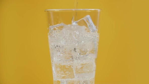 Pouring Transparent Fizzy Drink. Glass Full of Cold Sparkling Water with Ice Cubes Yellow Background