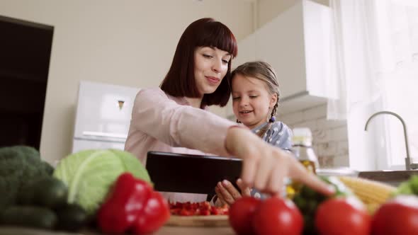 Happy Caucasian Young Daughter and Her Young Mother Having Online Cooking Class on Tablet