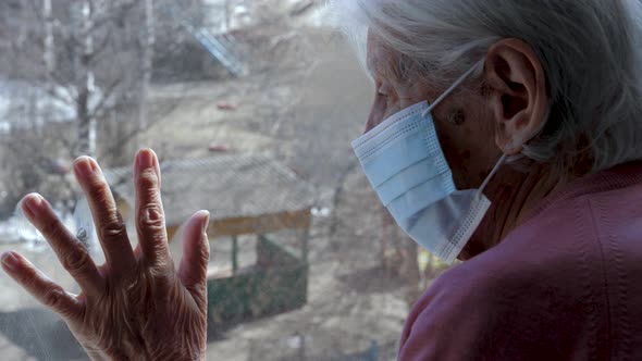 Old Woman In Medical Mask On Face Looks Out At Street With Her Hand On Glass
