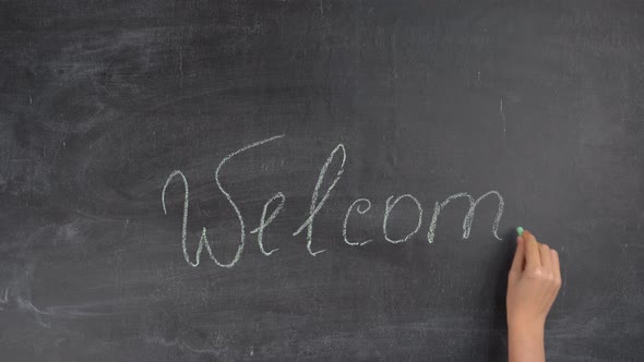 A woman's hand writes the word WELCOME in green chalk on a black chalk board