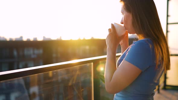 Woman with a Cup of Coffee Standing on the Balcony and Admire the Sunset