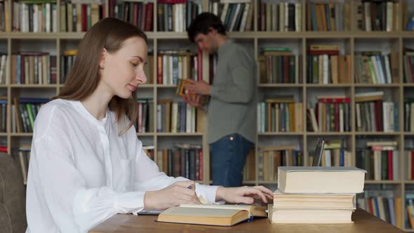 Young Woman Sitting at Table with Books and Laptop in University Library and Studying