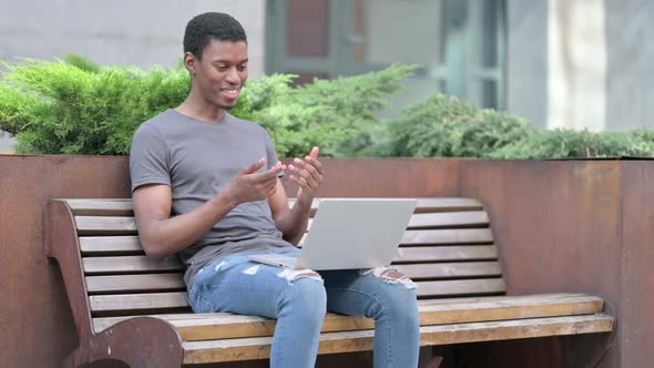 Cheerful Young African Man Doing Video Call on Laptop on Bench 