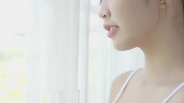 Young asian woman waking up and drinking glass of water with fresh and looking windows.