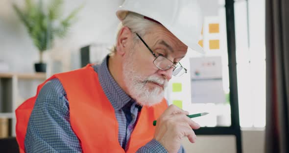 Mature Bearded Architect in Helmet and Vest Working with Blueprint