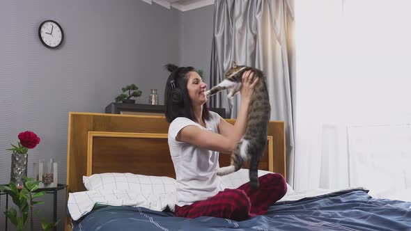 woman in headphones listening music on bed in bedroom and playing with cat