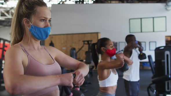 Two fit caucasian women and fit african american man wearing face masks exercising using kettlebell