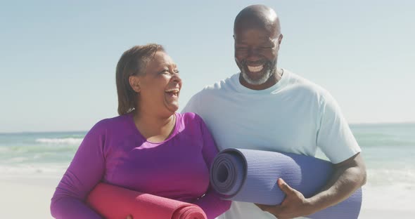 Portrait of happy senior african american couple holding yoga mats on sunny beach