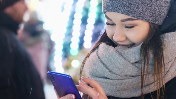 Close-up of Young Woman Uses the Mobile App in the Snowy Evening, Christmas and New Year