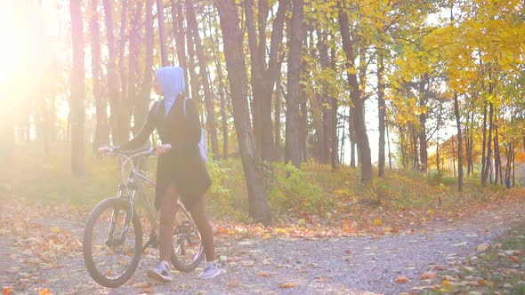 Muslim Woman in a Hijab and with a Backpack with a Bicycle Goes Through a Sunny Autumn Park Sun