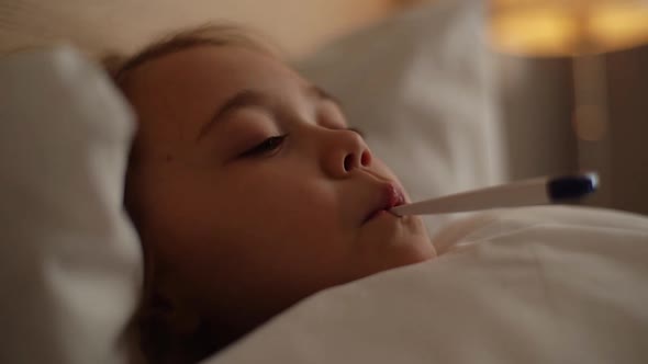 Closeup Face of Little Sick Girl with High Temperature Lying in Bed with Thermometer in Her Mouth in