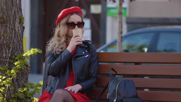 Young Beautiful Woman in a Red Beret and Dress Black Glasses and a Braid is Sitting on a Park Bench