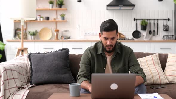 Cheerful Amazed Arabian or Indian Guy Freelancer Sits on a Sofa in Living Room Looks at Laptop