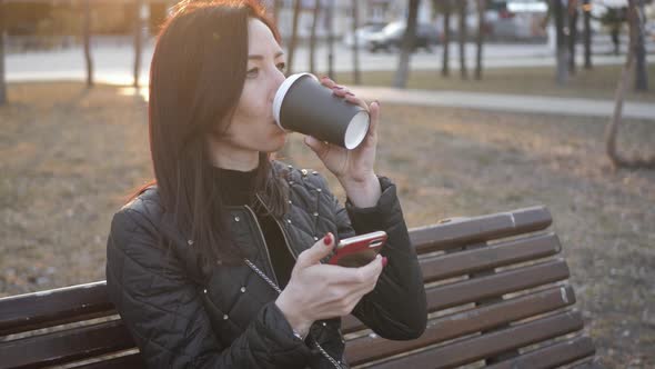 Beautiful Woman Use Phone Sitting on a Park Bench with Coffee Cup.