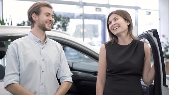 Happy Young Couple Standing at New Car in Dealership Talking, Looking at Camera and Smiling