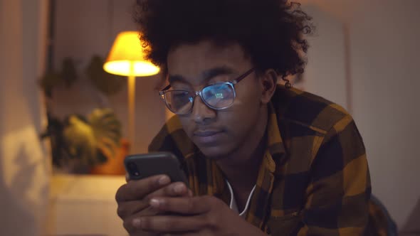 Young Black Man Using Mobile Phone Browsing Internet Lying in Bed at Home