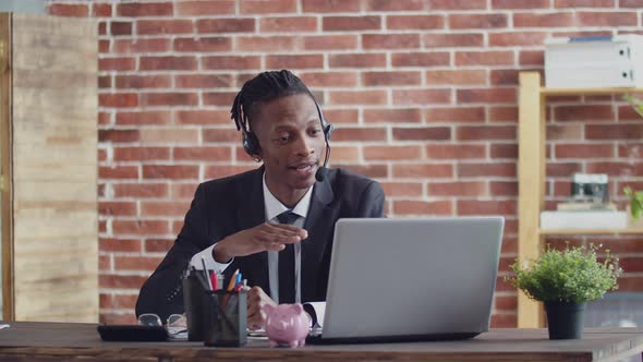 Black Man in a Suit and Headset Speaks Via Video Link Using a Laptop in Office at the Table