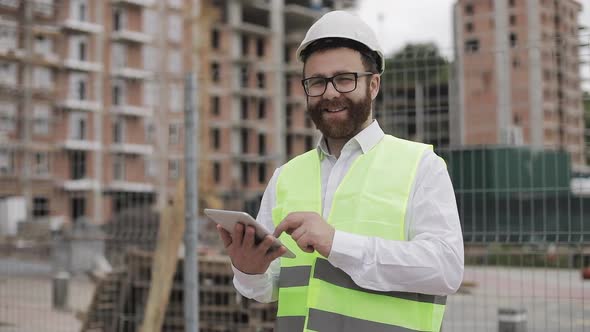 Portrait of a Successful Young Businessman, Wearing a White Helmet, in a Suit Working with Tablet