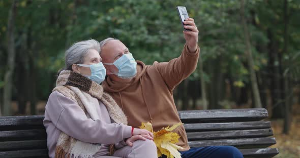 Adult Couple in Medical Masks, Sits on a Bench in a Forest Park and Take a Selfie Video on a