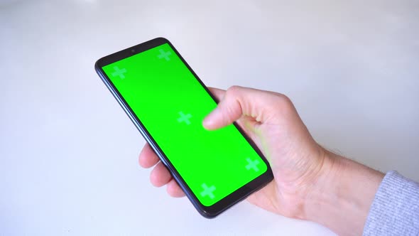 close-up. man scrolls through green chromakey screen, holding his thumb on smartphone one hand over