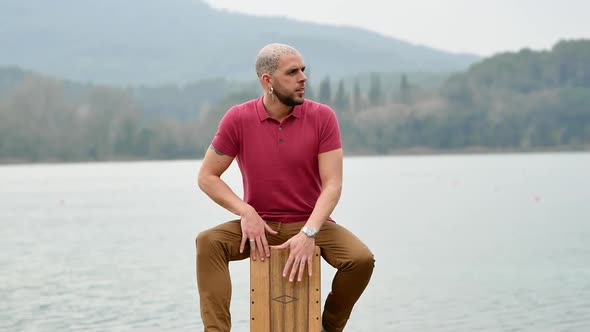 Pensive musician playing cajon in nature against lake and mountains