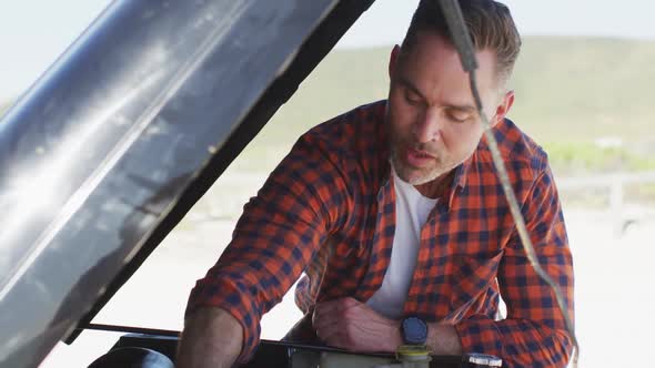 Happy caucasian man repairing broken down car with open bonnet on sunny day at the beach