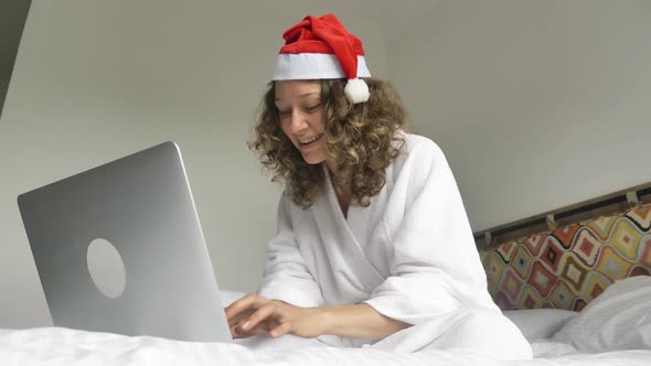 A Happy Businesswoman in a White Coat and a Santa Claus Hat is Texting with Friends Online on a