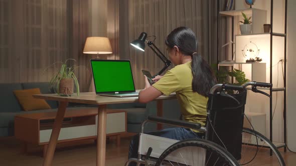 Asian Kid Girl Sitting In A Wheelchair While Using Laptop Green Screen And Use Mobile Phone At Home