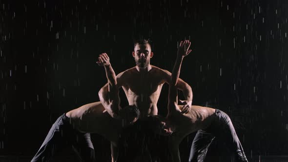 A Group of Three Acrobats Perform Difficult Balancing Tricks in the Rain. Men Stand Against Black