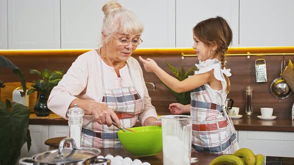 Grandmother is Whipping Eggs and Her Little Granddaughter is Staining Grannys Nose in Flour They