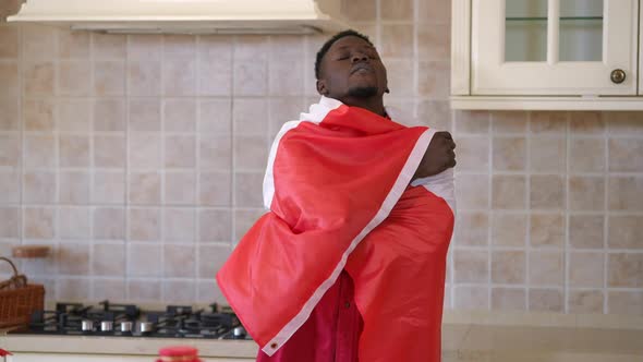 Young Happy African American Man Wrapping in Canadian Flag Standing in Kitchen at Home Looking at