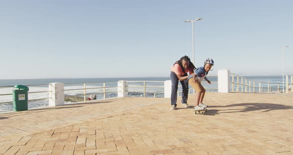 Video of happy african american father learning son how to skateboard on promenade