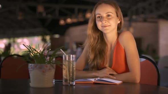 Blonde Girl Sits at Table with Juice Glass in Cafe