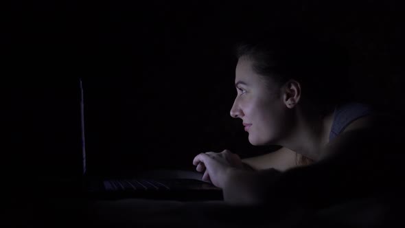 Attractive Beautiful Young Woman Lying on the Bed Using a Laptop in a Dark Room 