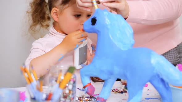 Step by step. Mother and daughter decorating paper mache unicorn with jewels and paper flowers.