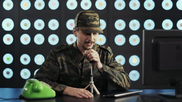 Guy in Camouflage Uniform at Desk with PC Speaks Via Paging Microphone in Hurry