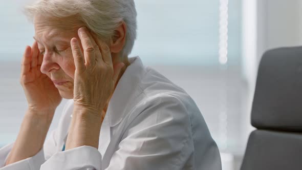 Tired grey haired woman doctor in robe massages temples suffering from headache