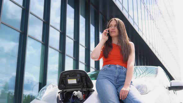 Attractive Woman Talking on Phone While Charging Electric Car Near the Shopping Center