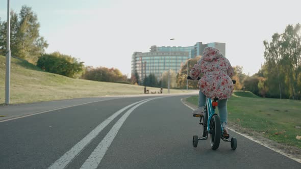 Little Girl Rides a Tricycle Bike in the City Park at Sunset in Sunny Autumn