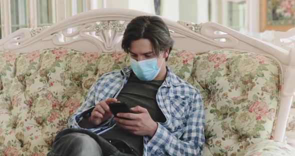 Young Man with Medical Mask on Face Using Smartphone When Resting on Sofa