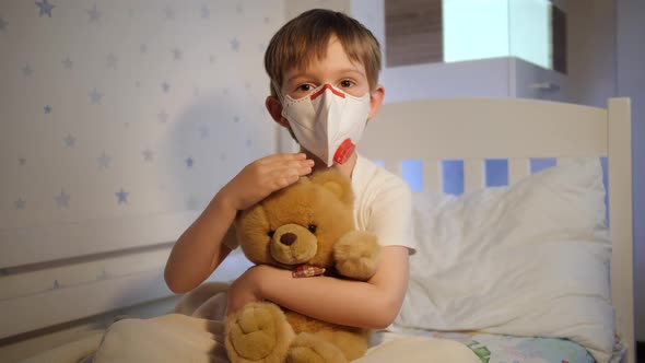 Cute Little Boy Wearing Protective Medical Mask Holding and Stroking His Teddy Bear in Bed at Night