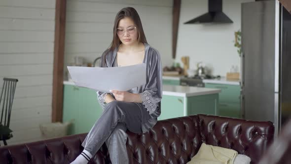 Wide Shot of Serious Confident Asian Young Woman Analyzing Business Plan Sitting on Couch Indoors