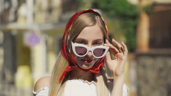 Portrait of Cute Young Woman in Sunglasses Looking at the Camera, Posing with Red Shawl on Her Head