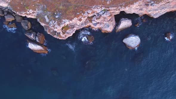 Top down aerial view of waves splash against rocky seashore, background. Flight over high cliffs of