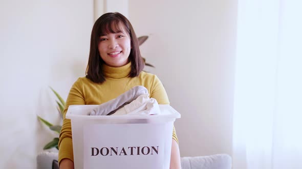 Portrait of young asian woman looking at camera holding box with clothes for donation.