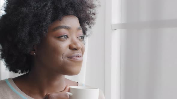 Calm Smiling Dreamy African Young Woman Enjoys Good Morning Holding Cup Looking at Window