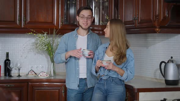 Joyful Millennial Boyfriend Talking with Girlfriend Drinking Morning Coffee Standing in Kitchen