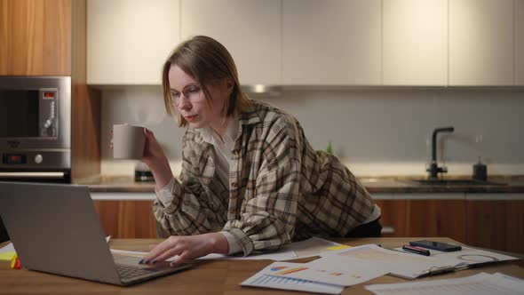 A Young Beautiful Business Woman Works From Home with a Cup of Coffee and Looks Into a Laptop Screen