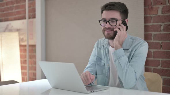 Young Male Designer Talking on Smartphone and Working on Laptop